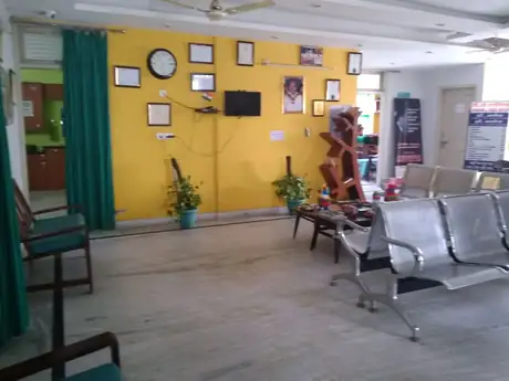 The clinic's waiting area, with a row of chairs, a yellow wall hung with certificates and a small photograph, two potted plants and a wall clock.